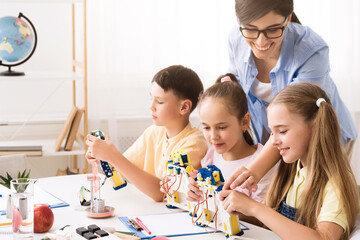 Aufkleber - A group of elementary school students are working on a robotics project in a classroom setting. They are building robots with the help of their teacher, who is smiling and guiding them.