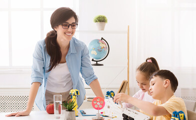 Poster - Woman teacher wearing glasses is smiling as she supervises two children building a robot in a classroom. The children are working together on the project, and the teacher is providing guidance