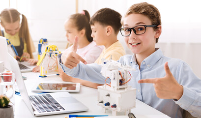 Poster - A young boy sits in a classroom setting with other students. He is smiling and pointing at a small robot he has built, demonstrating his learning and enthusiasm for STEM education.