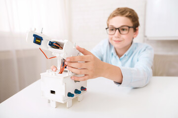Poster - A young boy wearing glasses is seen at a desk, focused on assembling a white robotic arm model. He carefully aligns the components, demonstrating his interest in robotics and STEM learning.