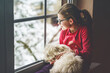 © Irina Schmidt - Little girl sitting by window with her pet dog Maltese at home. Happy child and cute puppy looking out on winter snow landscape. Love, friendship, family animal.