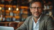 © JoxyAimages - A man with glasses and a stylish blazer smiles as he works on his laptop in a warmly lit cafe with a bookshelf background, capturing a friendly and productive atmosphere.