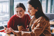 © BullRun - Two female friends enjoying meeting at coffee shop watching positive video blog using smartphone device, hipster girls casual dressed communicated in social networks with colleague from university
