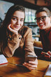 © BullRun - Portrait of positive female friends looking at camera while installing application for chatting using cafeteria wireless internet together, two happy hipster girls using smartphone devices indoors