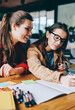 © BullRun - Positive smiling female student designers spending time together for working on startup project, young charming woman in spectacles drawing sketches sitting near happy friend at cafeteria table