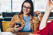 © BullRun - Selective focus on positive female blogger using smartphone device for creating publication sitting at table with laptop computer connected to wifi while friend on front making telephone call