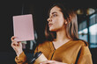 © BullRun - Serious hipster girl looking away and pondering on new best seller sitting on chair at coffee shop, thoughtful woman holding textbook for education and thinking about literature hobby indoors