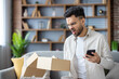 © Tetiana - Frustrated man opening delivery box at home while holding smartphone, unhappy with order