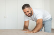 © Prostock-studio - A man in a white t-shirt and grey sweatpants is smiling as he makes his bed in a bedroom. His arms are outstretched, and he is smoothing the sheets down.