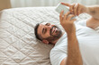 © Prostock-studio - A man with a beard lies on a bed with a white duvet and looks at his smartphone, smiling. He is wearing a white t-shirt, top view