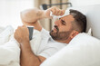 © Prostock-studio - A man lies in bed with a tissue in his hand, wiping his forehead. He appears to be sick or unwell, as he holds a mug and has a tired expression on his face. He is covered by white bedsheets