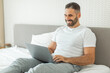 © Prostock-studio - A man with a beard is sitting on a bed and using a laptop. He is wearing grey sweatpants and a white t-shirt. He is smiling and looking at the laptop screen. The bed is made with white sheets