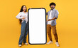 © Prostock-studio - Cool mobile offer. Happy indian couple pointing at big cellphone with white screen for mockup, demonstrating free space for phone application or website design, yellow background
