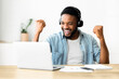 © Prostock-studio - A man in a blue shirt pumps his fists in joy while wearing headphones at his desk, celebrating a success on his laptop.