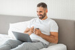 © Prostock-studio - A man sits in bed, using a laptop computer and holding a cup of coffee. He is smiling as he looks at the laptop screen. The bed has white sheets and pillows.
