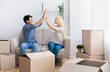 © Prostock-studio - Successful Relocation. Young Couple Giving High-Five Sitting On Sofa Among Moving Boxes In New Apartment. Free Space