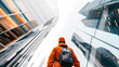 © Natalia - Low-angle view of a person in a hard hat and orange jacket looking up at modern glass buildings.
