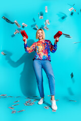 Poster - Full length vertical photo of overjoyed cool girl dressed print shirt jeans shooting money with guns isolated on blue color background