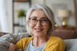 © john - Smiling middle aged mature grey haired woman looking at camera, happy old lady in glasses posing at home indoor, positive single senior retired female sitting on sofa in living room headshot portrait