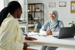 © pressmaster - Young cheerful general practitioner in hijab looking at pregnant African American patient during medical consultation in office
