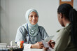© pressmaster - Young smiling female doctor in hijab using tablet and consulting African American patient filling in medical form while visiting clinician