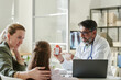 © pressmaster - Confident male pediatrician pointing at bottle of pills or vitamins in his hand and looking at cute little patient visiting him with her mother