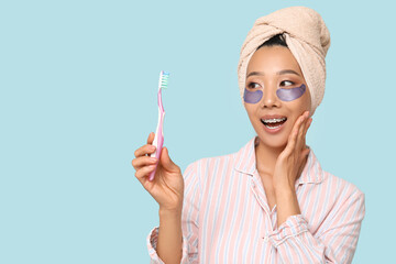 Poster - Young Asian woman with under-eye patches and toothbrush on blue background, closeup