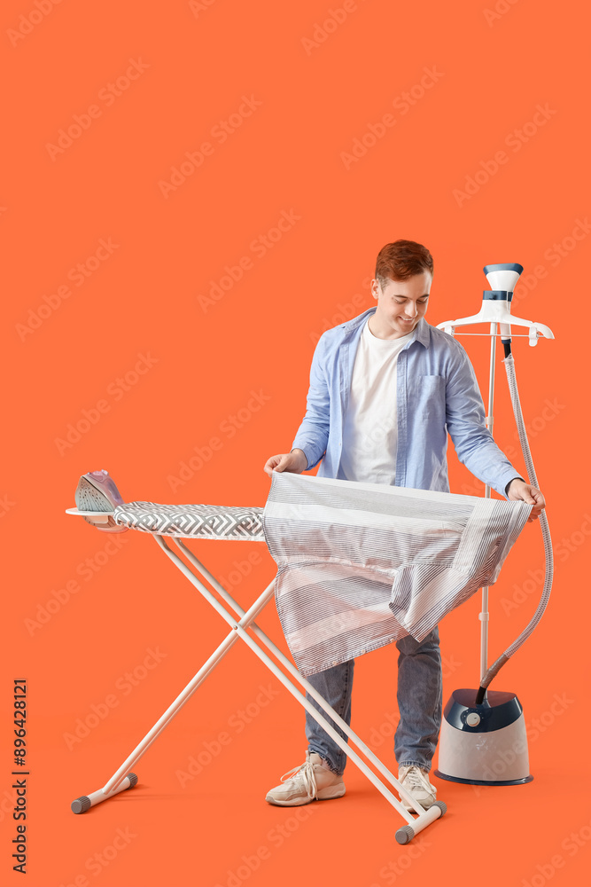 Handsome young man ironing clothes on orange background