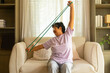 © CandyRetriever  - Happy Asian senior mature woman enjoy healthy lifestyle resistance band training exercise for stretching body muscle shoulder and arm on sofa in living room. Elderly self-care and home fitness concept