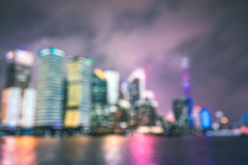 Canvas Print - A blurred out nighttime view of a city skyline, lit up by streetlights, with a body of water in the foreground and a cloudy sky in the background.