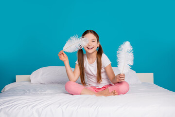 Poster - Photo of pretty cheerful girl sitting cozy bed bedroom big feather isolated on blue color background