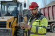© Centric  - A construction engineer in a red hard hat and safety goggles, using a tablet at a construction site with heavy machinery in the background