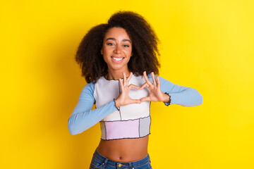 Poster - Photo of adorable positive girl with perming coiffure dressed stylish shirt showing heart symbol isolated on yellow color background