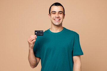 Canvas Print - Young happy rich middle eastern man wear blue t-shirt casual clothes hold in hand mock up of credit bank card look camera isolated on plain pastel beige background studio portrait. Lifestyle concept.