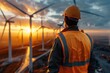 © Nena Ai - A worker in a hard hat and orange vest stands on a site with multiple wind turbines in the background, observing the renewable energy machinery as the sun sets.