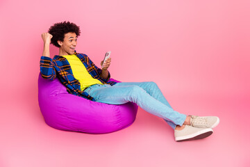 Poster - Photo of cheerful glad nice man wearing plaid stylish clothes sitting soft pouf fist yes isolated on pink color background