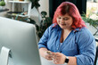 © LIGHTFIELD STUDIOS - A plus size businesswoman with pink hair checks her phone while sitting in front of a computer.