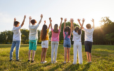 Wall Mural - Back view group of children standing in a line on a green grass lawn in the summer park in the morning, looking up at the sky and raising their hands to the sun. Nature, summer, childhood concept