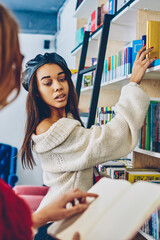 Two clever female students making research for project in school library, young woman reading literature while teenage friend standing near bookshelves and choosing best seller for leisure indoors