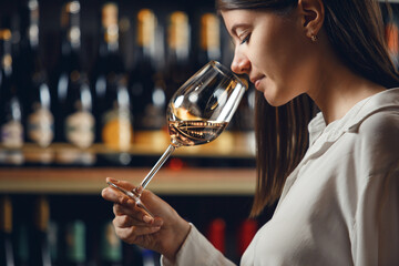 Professional woman sommelier smells white wine from glass, standing in cellar against shelf various alcoholic beverage background.