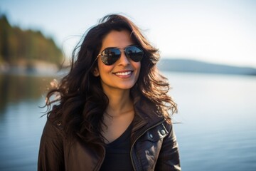 Wall Mural - Portrait of a smiling indian woman in her 30s wearing a trendy sunglasses in serene lakeside view