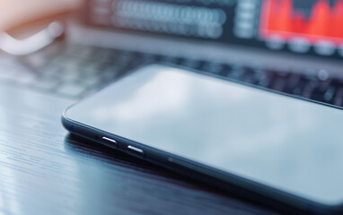 Poster - A smartphone placed on a wooden desk in front of a laptop showing financial charts. The scene is blurred, focusing on the sleek phone.