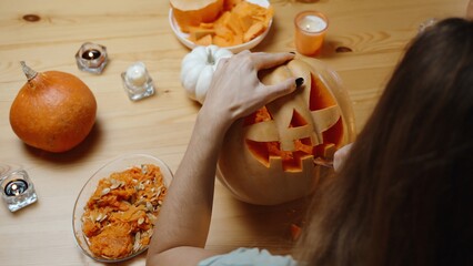 Wall Mural - On a Wooden Table in Warm Tones of a Cozy Home Setting, a Woman Carves a Jack-O'-Lantern from a Pumpkin. Candles and Pumpkin Insides on the Table, View from Behind and Above.