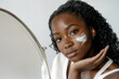 © Sachin - Young African American woman applying face cream, beauty skincare routine with mirror reflection