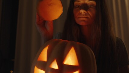 Wall Mural - In the Dark Room, the Woman Peeking Inside the Jack-o'-Lantern, Her Face Illuminated by Bright Warm Light from the Flame. She Closes It with the Lid. Slow Motion.