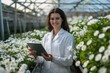 © Ольга Лукьяненко - Portrait of young smiling female agronomy expert in lab coat using tablet standing between flower beds