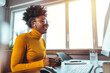 © Dragana Gordic - Smiling mid adult Black businesswoman sits at desk with computer, exudes confidence in casual attire, engaging in work from a sunlit contemporary home office.