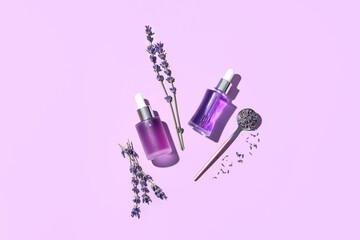 Bottles of lavender essential oil, teaspoon and flowers on lilac background