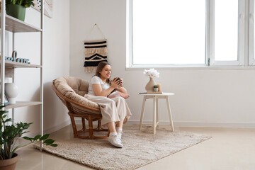 Canvas Print - Beautiful young woman sitting in armchair with cup of tea in living room