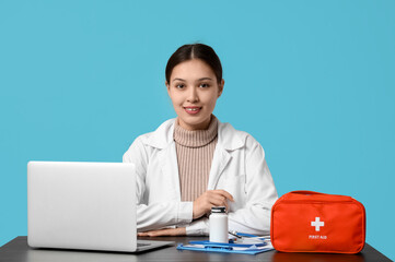 Poster - Asian female doctor with first aid kit, jar of pills and modern laptop at table on blue background